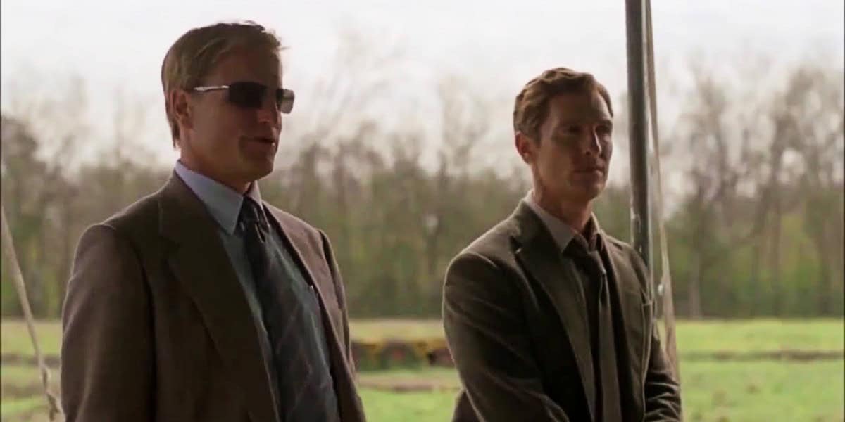 Two men wearing suits and ties stand outdoors under a canopy. One man is wearing sunglasses. The background features a field and trees under an overcast sky.