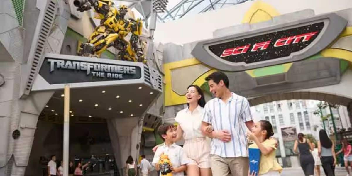 A family of four smiles and walks in an amusement park, near an entrance labeled "Transformers The Ride." The area has a futuristic, sci-fi theme with robot decorations and a large "Sci-Fi City" sign overhead.