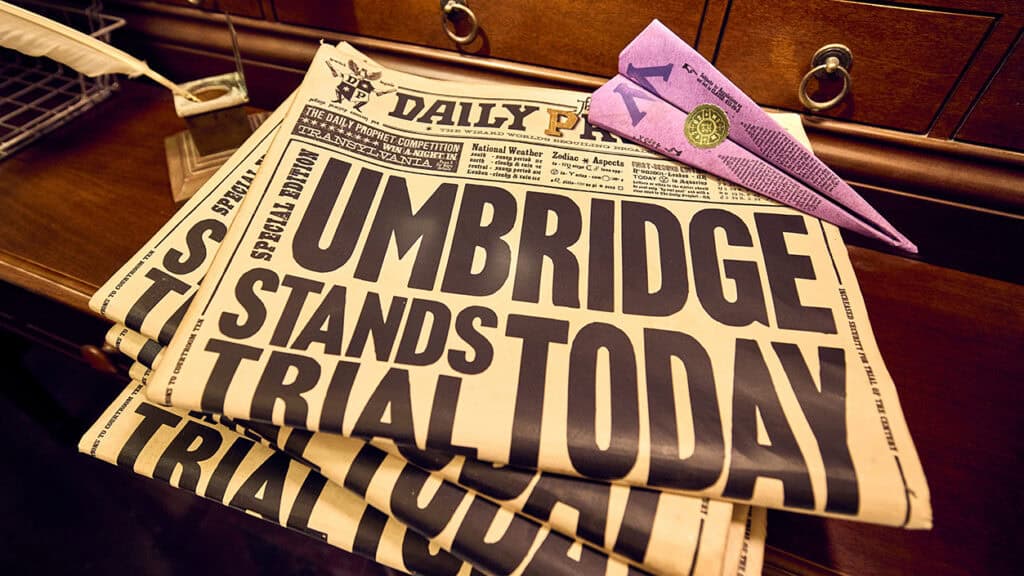 A stack of newspapers with the headline "UMBRIDGE STANDS TRIAL TODAY" in bold letters. A purple envelope with a seal and folded into a paper airplane shape rests on the top newspaper. The background features a wooden surface and drawers.