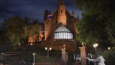 A gothic-style haunted mansion at night, reminiscent of Disneyland's eerie charm, is illuminated by subtle lighting. A vintage carriage is parked outside, and a ghostly figure in transparent clothing enhances the spooky atmosphere. Majestic green trees frame this chilling scene.