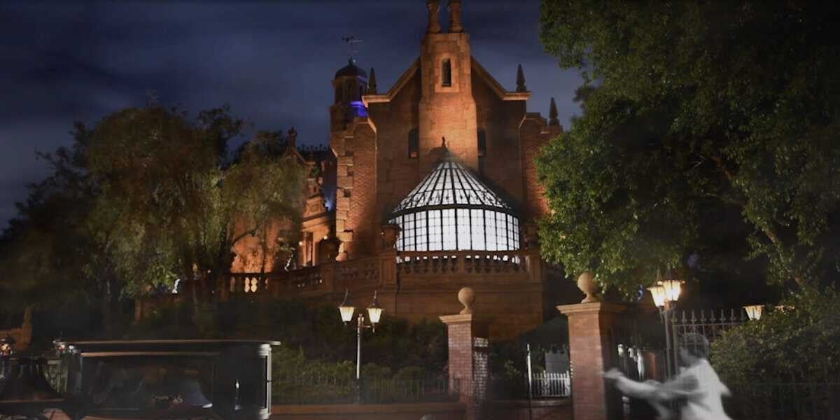 A gothic-style haunted mansion at night, reminiscent of Disneyland's eerie charm, is illuminated by subtle lighting. A vintage carriage is parked outside, and a ghostly figure in transparent clothing enhances the spooky atmosphere. Majestic green trees frame this chilling scene.