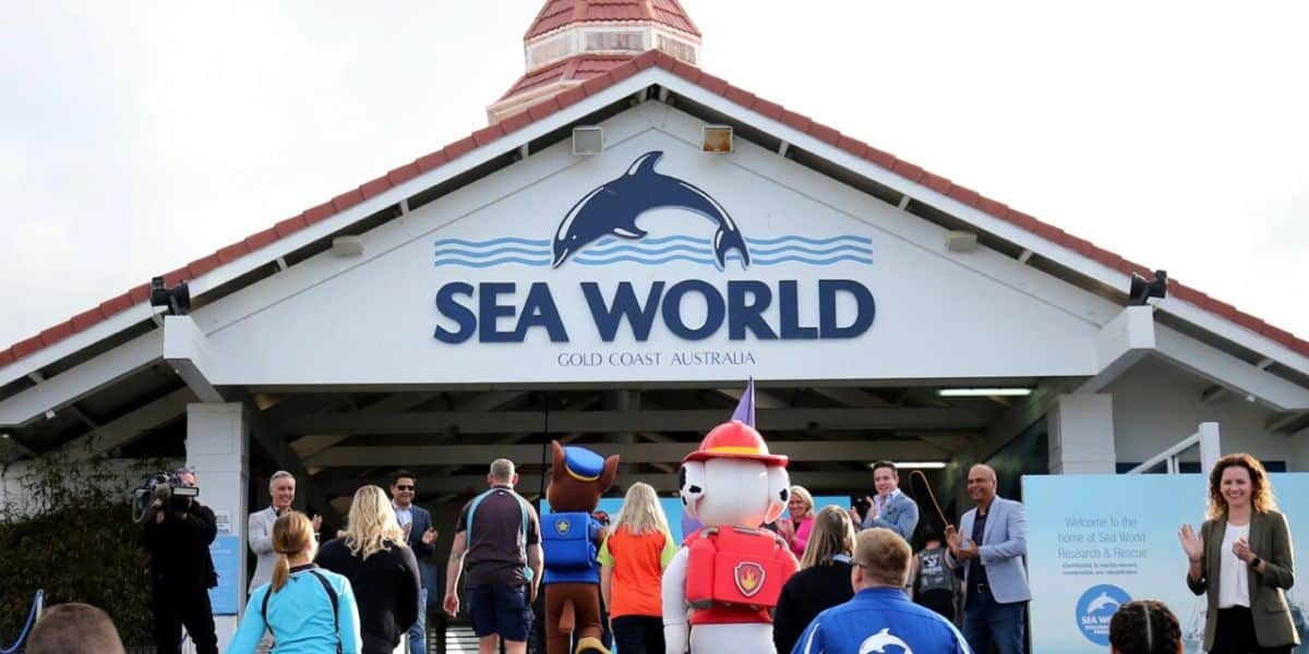 A group of people, including characters in costumes, are entering the Sea World entrance in Gold Coast, Australia. The entrance features the logo of a dolphin above the words "Sea World.