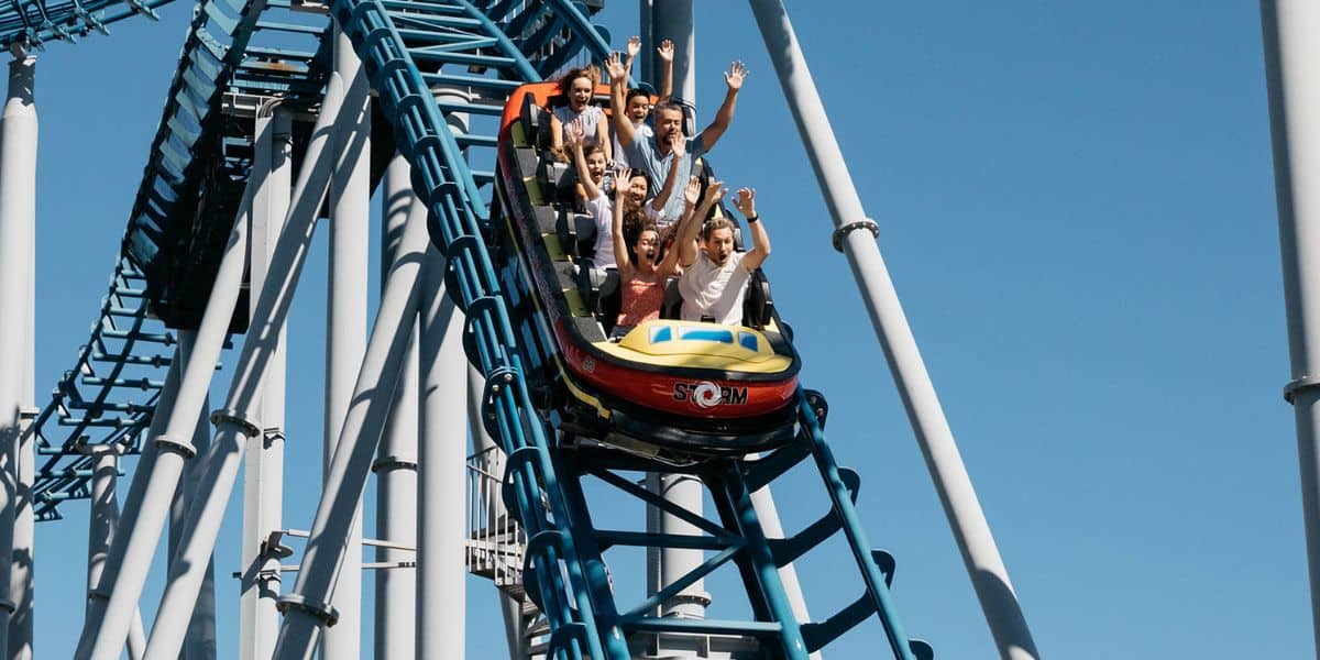 A group of excited people with raised arms enjoy a thrilling ride on a roller coaster, surrounded by blue metal tracks against a clear blue sky.