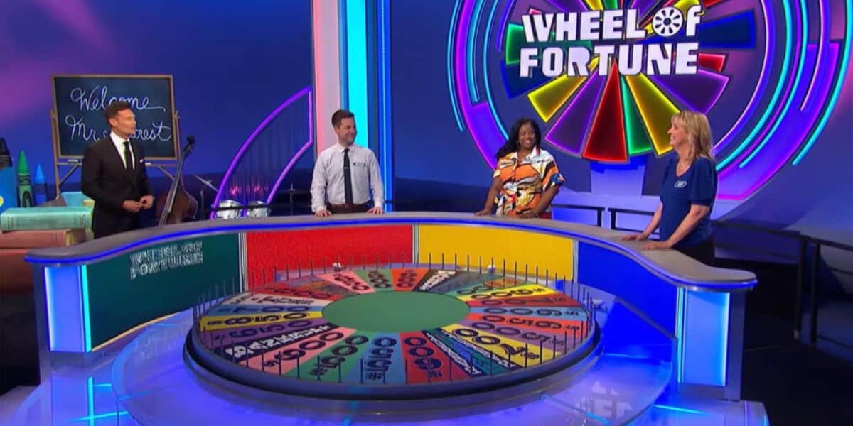 Four people stand around the Wheel of Fortune game show set. The colorful wheel is visible in front of them, and the neon-lit logo is displayed in the background.