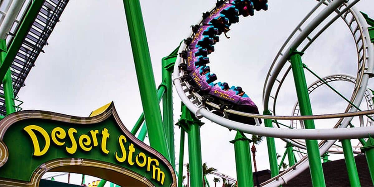 A vibrant roller coaster called "Desert Storm" twists and turns on bright green tracks under a cloudy sky. The purple coaster train is filled with excited riders. The entrance sign with the ride's name is prominently visible in the foreground.