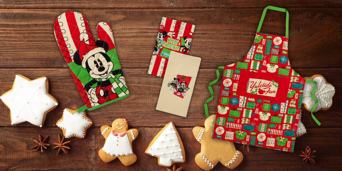 A festive holiday scene displays a red and green apron and oven mitt with cartoon character designs, surrounded by decorated cookies shaped like stars, a Christmas tree, and gingerbread people on a wooden surface.