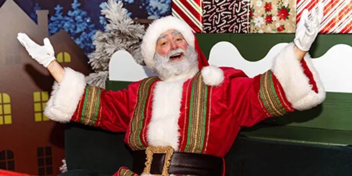 Santa Claus, in a festive red and gold-trimmed suit, sits joyfully with arms outstretched amidst SeaWorld's enchanting Christmas setting. Wrapped presents fill the background, enhancing the holiday magic.