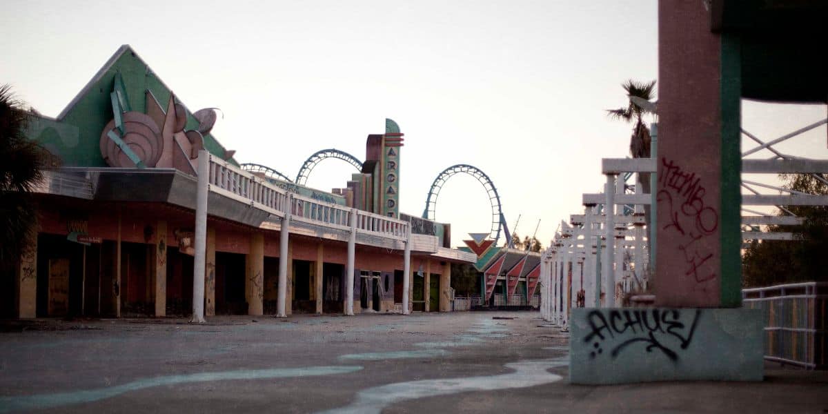 Abandoned amusement park with faded, graffiti-covered buildings and a visible roller coaster in the background. The setting appears deserted, with palm trees and empty walkways under an overcast sky.