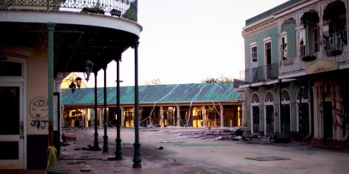 A deserted street with historic buildings, featuring ornate ironwork and arched windows. Debris and disrepair are evident, suggesting neglect or aftermath of a disaster. The scene is calm with warm light casting long shadows.