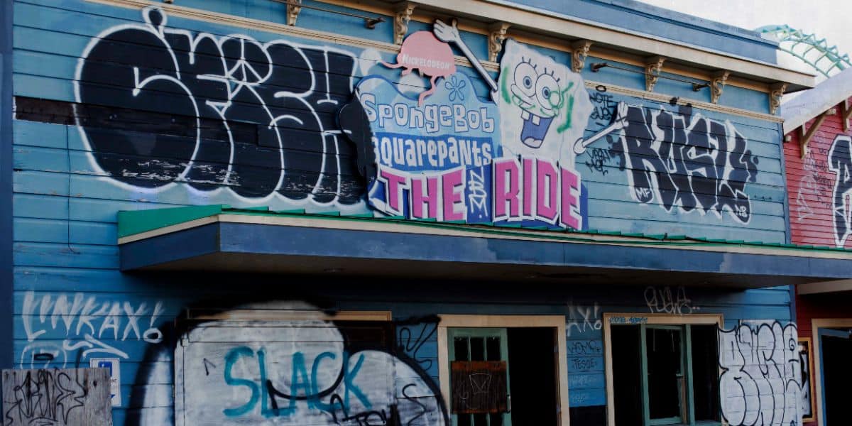 A closed amusement ride with a "SpongeBob SquarePants THE RIDE" sign above the entrance. The building is covered in graffiti, featuring multiple tags and artistic designs on the blue wooden facade. The entrance is boarded up.