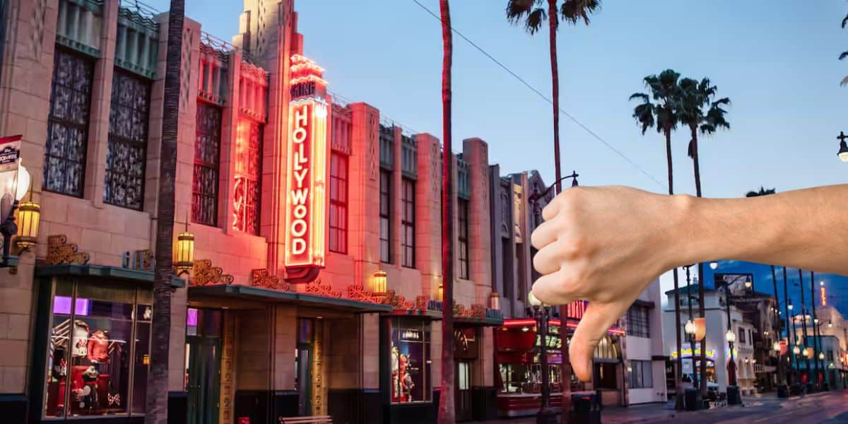 A neon sign reading "Hollywood" glows in the evening light on a building's façade. The street is lined with palm trees. In the foreground, a hand gives a thumbs-down gesture.
