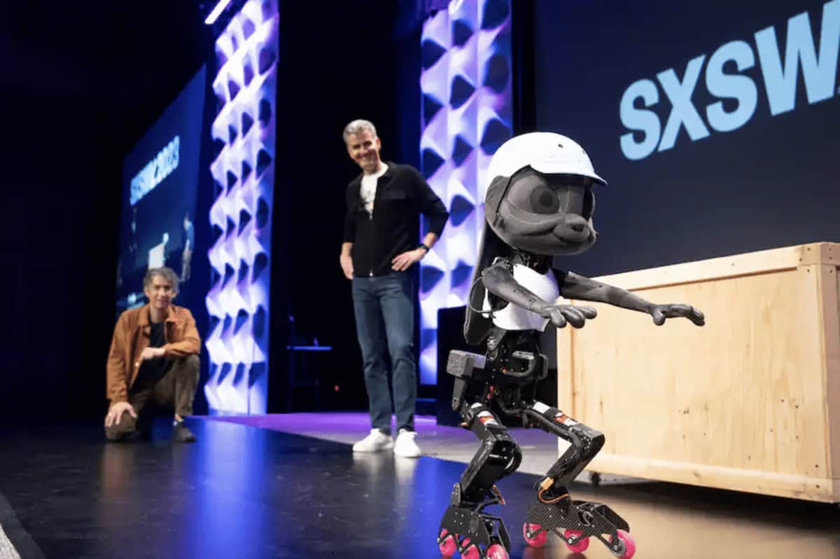 A small humanoid robot wearing roller skates and a helmet dazzles on stage at SXSW, reminiscent of undelivered Disney projects. Two people in the background observe its performance, smiling as they watch the demonstration.