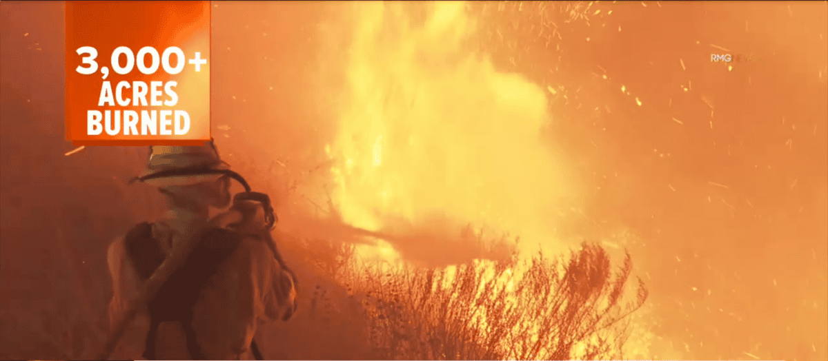 A firefighter stands in front of a large, intense wildfire. The sky is filled with orange and yellow flames. Text on the image reads "3,000+ acres burned.