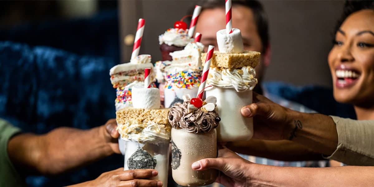 A group of people at the Toothsome Chocolate Emporium in Hollywood are toasting with extravagant milkshakes topped with whipped cream, sprinkles, marshmallows, and cherries. Red and white straws add a festive touch, and everyone appears to be enjoying themselves.