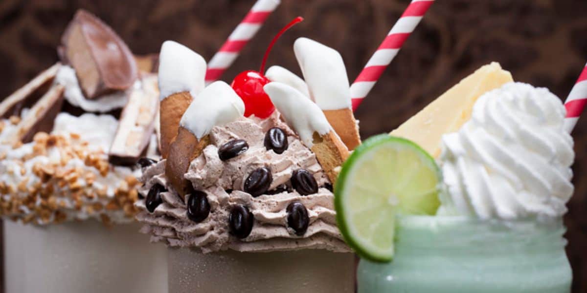 Three milkshakes with elaborate toppings: One with nuts and chocolate, another with whipped cream, cookies, coffee beans, and a cherry, and the third with whipped cream, a lime slice, and a cheese slice, each garnished with striped straws.