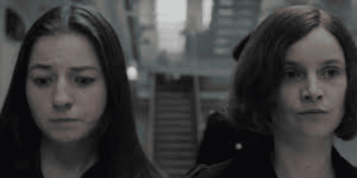 Two people with serious expressions stand side by side. One has long dark hair and the other has shoulder-length brown hair. A staircase and blurred background suggest an indoor setting.