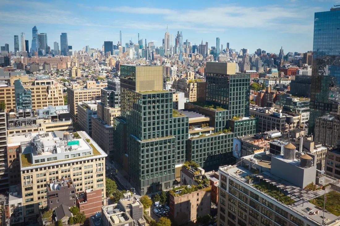 Aerial view of a cityscape featuring modern, green-roofed buildings in the foreground and an expansive skyline with skyscrapers under a clear blue sky in the background, showing the new Disney HQ in NYC.