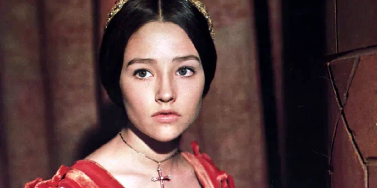 A young person with long dark hair, wearing a red and gold Renaissance-style dress and a cross necklace, looks to the side against a blurred background.