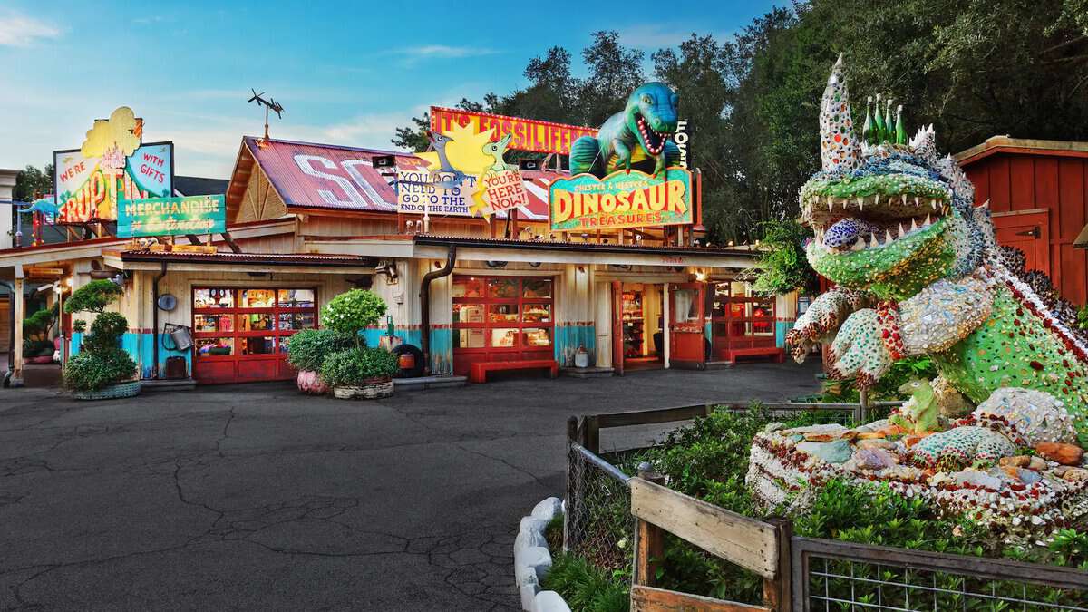 Hester and Chester Dinoland is a colorful, vintage-style roadside attraction featuring a building adorned with dinosaur-themed signs. A large, whimsical dinosaur sculpture made from assorted materials stands in a garden area. Lush greenery and a clear blue sky provide the perfect backdrop.