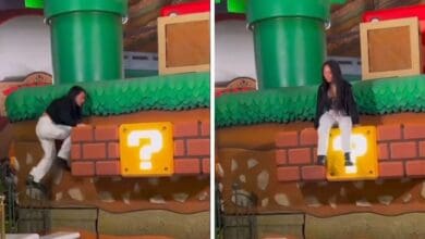 A person in a black jacket and white pants playfully climbs onto a large, yellow question block reminiscent of a video game at a themed park. The scene includes green pipes and pixelated bricks in the background.