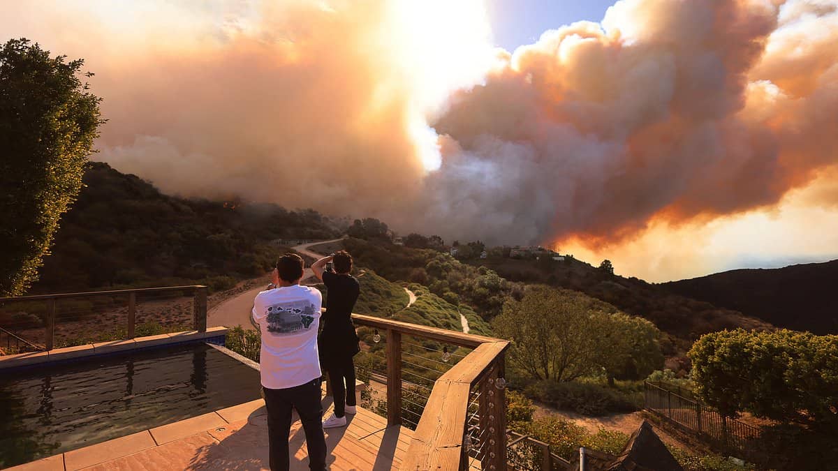 Two people stand on a deck overlooking a large wildfire in a hilly area. Thick smoke billows into the sky, partially obscuring the sun. A small pool is in the foreground, and dense vegetation surrounds the scene.