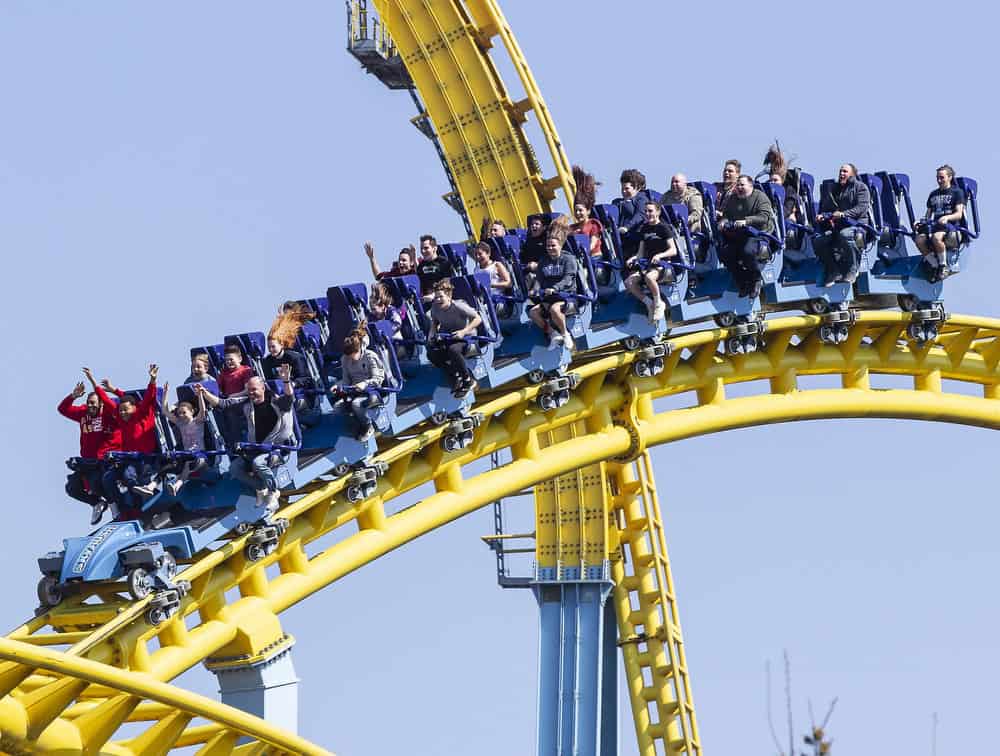 A group of people riding a roller coaster with yellow tracks and blue seats. The riders are excited, with some raising their arms and others with hair flying. The coaster twists against a clear blue sky at this theme park.