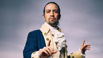 A person with long hair tied back is dressed in an 18th-century style outfit, featuring a blue coat, ivory waistcoat, and a white cravat. They gesture with one hand raised, set against a cloudy sky backdrop.