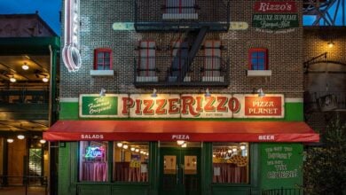 Front view of PizzeRizzo, a pizzeria with a brick facade and neon lights. The green and red signage reads "PizzeRizzo" and "Pizza Planet." The entrance features a red awning, tables, and chairs. The building is lit up against the evening sky.