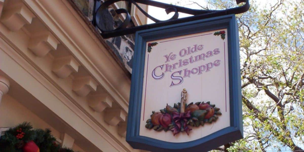 A hanging sign for "Ye Olde Christmas Shoppe" with decorative holly and fruit accents, set against a blue border. The building features white trim and festive garland. A tree with bare branches is visible in the background.