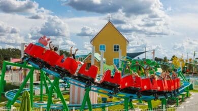 A bright red roller coaster twists through an amusement park with excited riders raising their arms. The track is green, and a colorful building stands in the background under a partly cloudy sky.
