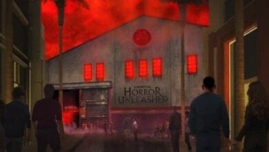 People walking toward a building with a red glow and smoke. The building has "Universal Horror Unleashed" written on it, with eerie red windows and palm trees. It is night, creating a spooky atmosphere.