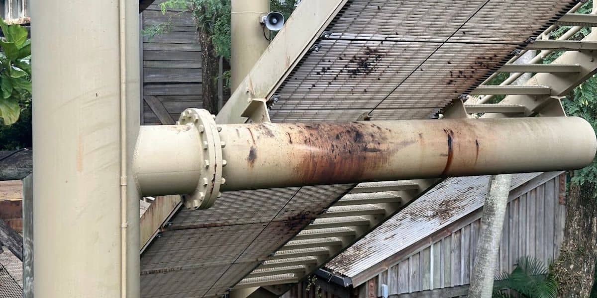 Metal pipe with rust spots running horizontally under an outdoor structure made of metal grates, supported by beams. There's a small security camera above the pipe. In the background, wooden buildings and greenery are visible.