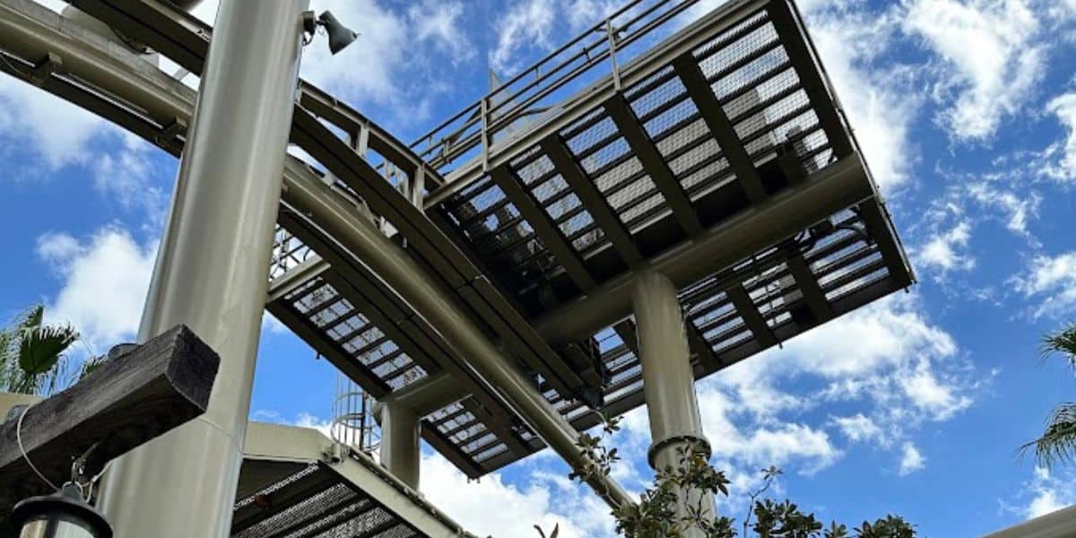 A view from below of a Universal roller coaster track against a blue sky with scattered white clouds. The metallic track is elevated and supported by large pillars, offering thrilling twists. Lush green trees are visible at the bottom of the image, enhancing the vibrant scene.