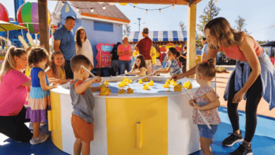 A group of children and adults gather around a colorful outdoor game table with rubber ducks. The scene is joyful and casual, with colorful structures and clear blue skies in the background.