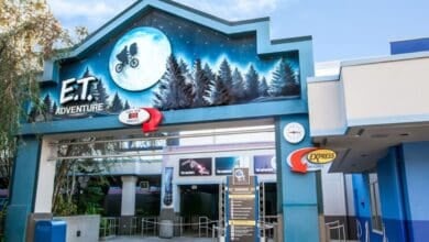 Entrance to the E.T. Adventure ride, featuring a large mural with E.T. silhouetted against a moon, bicycle flying through a forest. The building is blue with trees painted on. Signs for standby and Express entry are visible.