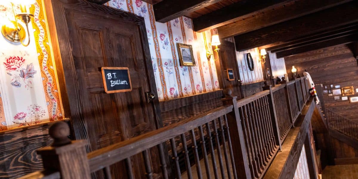 A rustic hallway with wooden walls and floral wallpaper. A door features a blackboard sign reading "B.T. Pullum." Wall sconces provide warm lighting, and framed pictures decorate the corridor. The wooden railing leads to an upper floor.