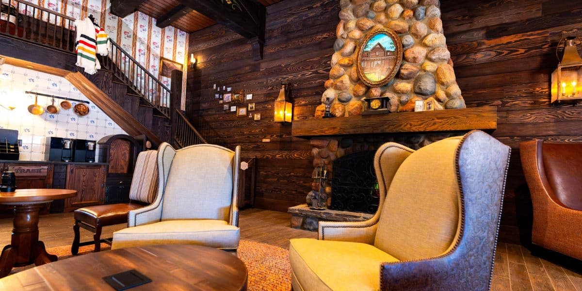 A cozy lodge interior with stone fireplace, wooden walls, and various armchairs around a coffee table. A staircase with patterned wallpaper leads upstairs. Warm lighting enhances the rustic atmosphere.