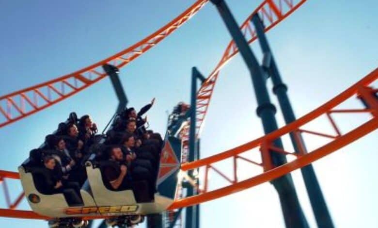 People ride a roller coaster with orange tracks under a clear blue sky. The car is white with "SPEED" written on the side, and passengers appear excited or thrilled as they experience the ride.