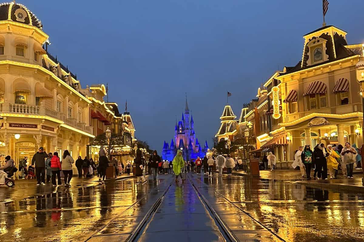 A rainy evening scene at Disney World, featuring a brightly lit castle in the distance. The wet street reflects lights from surrounding illuminated buildings.