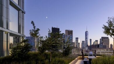 A rooftop garden with lush greenery overlooks a city skyline at dusk. The skyline features tall buildings, including one with a spire. Two people sit at a bench enjoying the view. The sky is clear with a visible crescent moon.