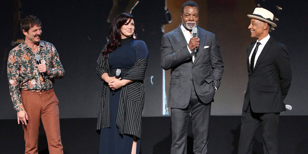 Four people stand on stage, each holding a microphone. The first wears a floral shirt and brown pants; the second, a navy dress with a striped overlay; the third, a gray suit; and the fourth, a black suit with a white shirt and hat. They are smiling and engaged in discussing progress on Gina Carano's lawsuit against Disney.