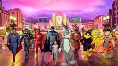 A child walks hand-in-hand with Batman, surrounded by people in superhero and cartoon character costumes, including Superman, Flash, Scooby-Doo, Bugs Bunny, and Tweety, in a brightly lit, colorful theme park street as a new Harry Potter is announced for WB Dubai.