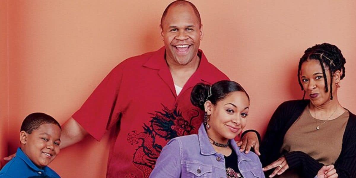 Four people pose in front of a peach-colored wall, smiling and looking cheerful. A young boy leans on a man in a red shirt, next to a young woman in a purple jacket, and a woman with braids stands beside them.