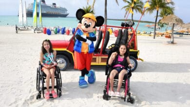 Two smiling girls in wheelchairs pose with Mickey Mouse on a sunny beach. Palm trees, a colorful jeep, sailboats, and a cruise ship are visible in the background.