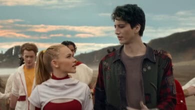 A blonde girl in a white and red outfit and a tall boy with dark hair wearing a green jacket walk together outdoors, looking at each other, with other people in the background. The sky is partly cloudy at sunset.