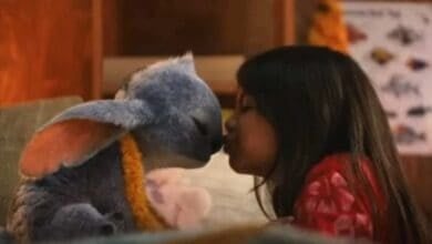 A young girl kisses a blue, plush-like creature with large ears and a flower lei. They are indoors, with shelves and a poster in the background.