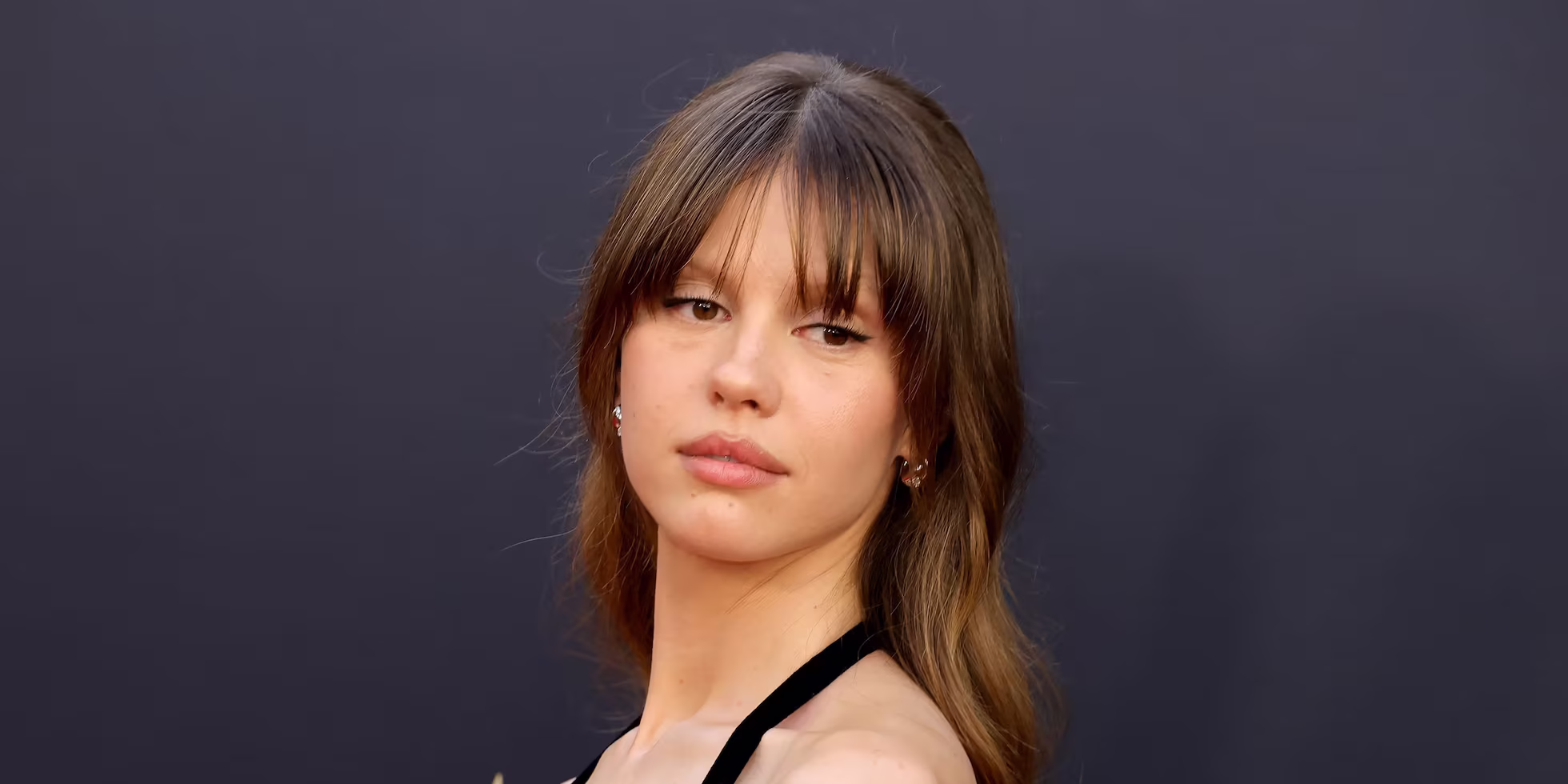 A woman with long brown hair and bangs, reminiscent of Mia Goth cast in Star Wars, wears a black leather dress with metal eyelet details, posing on a red carpet against a dark background.