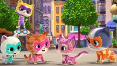 Four animated cats in colorful superhero costumes gather in a city square, while a fifth cat hovers above on a hoverboard. The playful scene, filled with vibrant buildings and green trees, captures the fun spirit of a Disney Jr adventure.