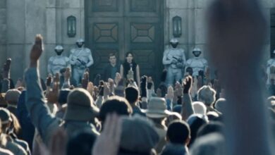 A crowd raises their hands in salute before a large building, where two people stand flanked by armored guards in white helmets and uniforms. The atmosphere is tense and orderly.