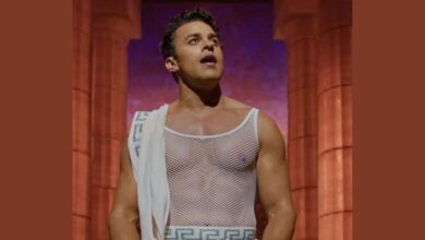 A man stands before reddish stone columns, dressed in a white mesh top and a Greek key-patterned drape. Channeling the Hercules Musical, he gazes upward with a surprised or amazed expression.